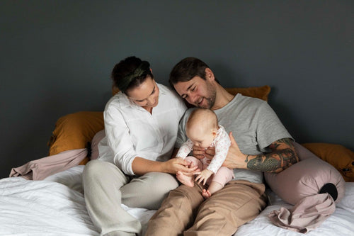 A baby and parents on a bed with a pregnancy pillow behind them